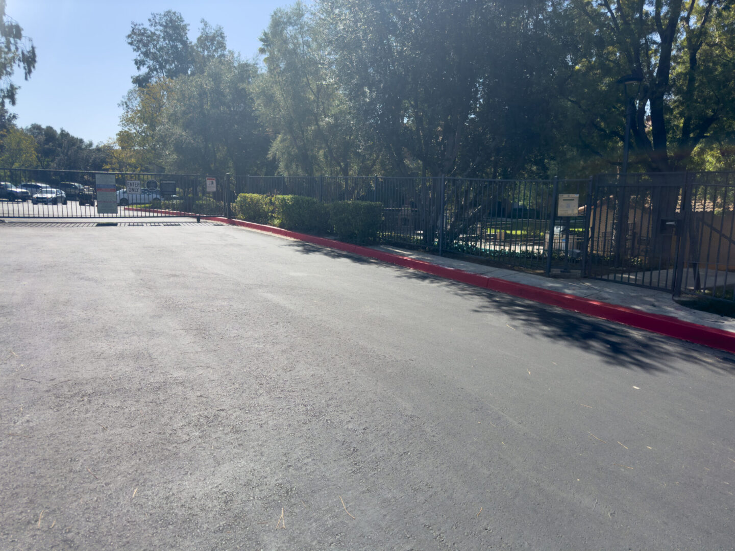 A newly paved road leads to a gated entrance surrounded by trees on a sunny day. A "No Parking" zone is marked along the curb with red paint.