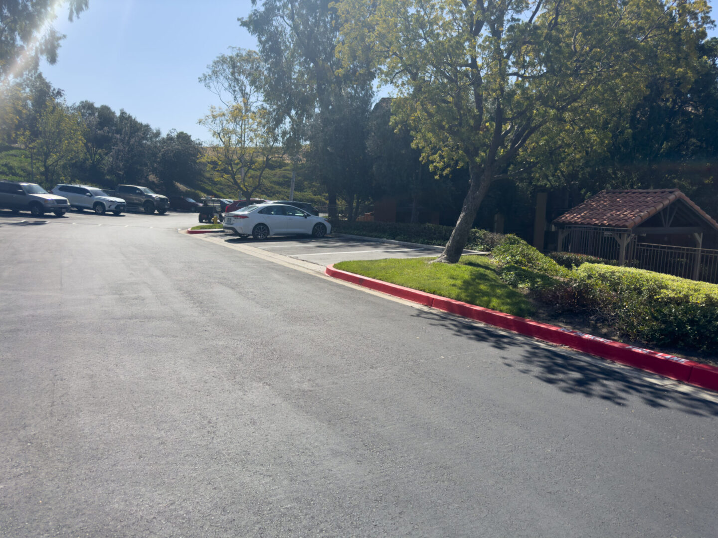 A sunny parking area with several cars parked. Trees and a small building with a red-tiled roof are in the background alongside a landscaped area.