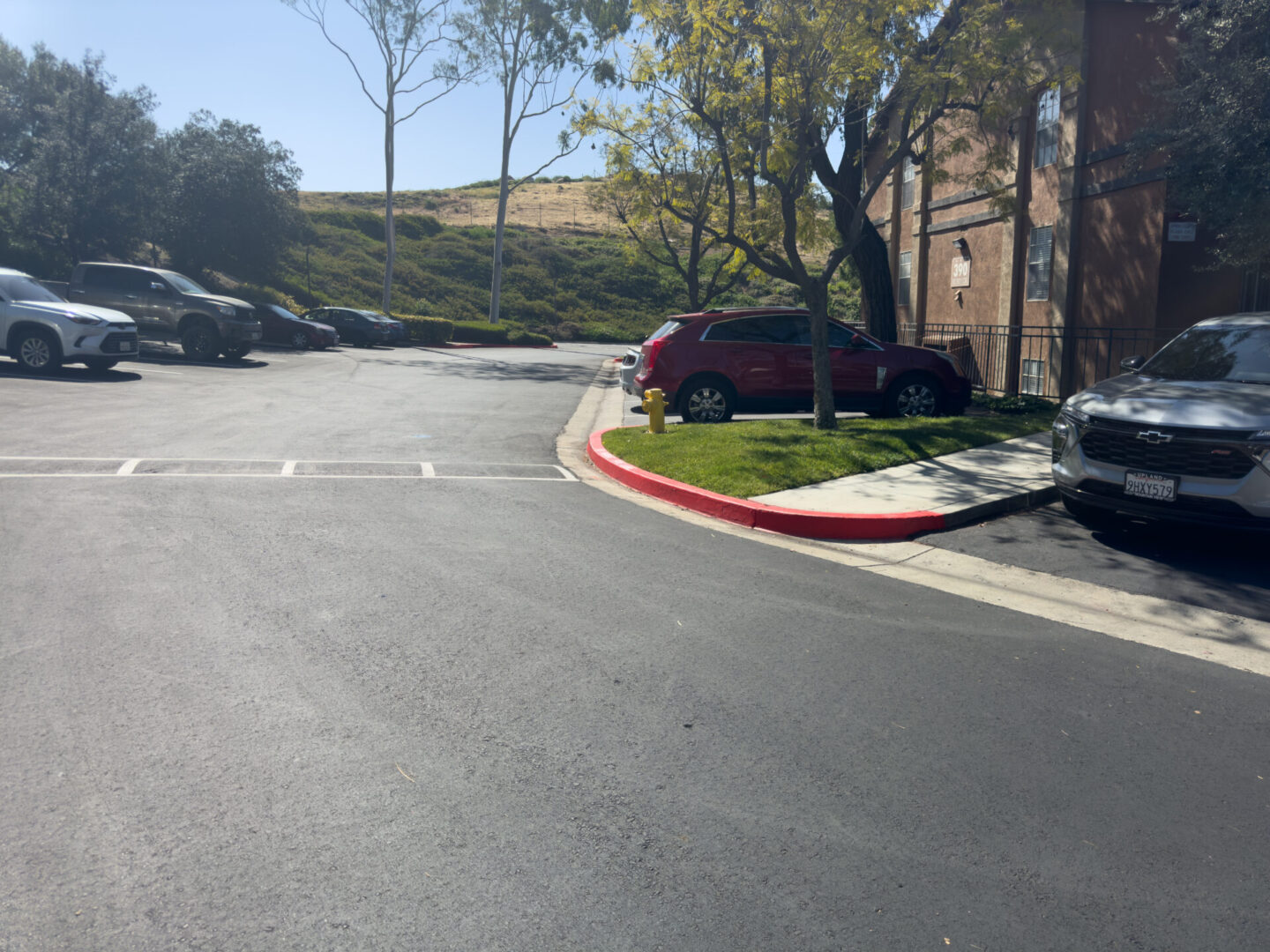 A parking lot with several parked cars, bordered by a red-painted curb, trees, and hilly grassy area in the background under a clear sky.