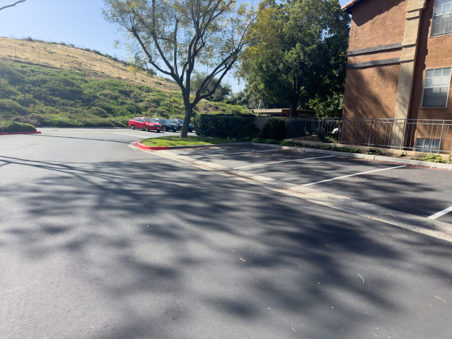 An empty parking lot next to a multi-story building with trees and a hill in the background, and a red car parked in the distance.