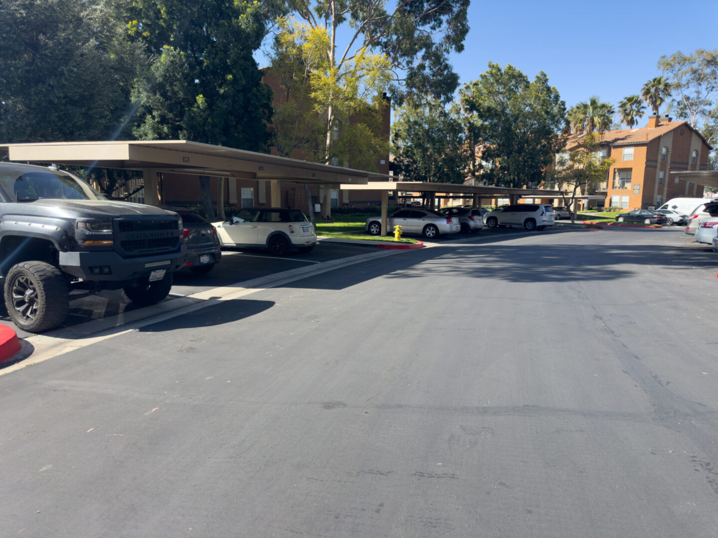 A parking lot with several cars parked under a covered parking area on a sunny day. Trees and residential buildings are visible in the background.