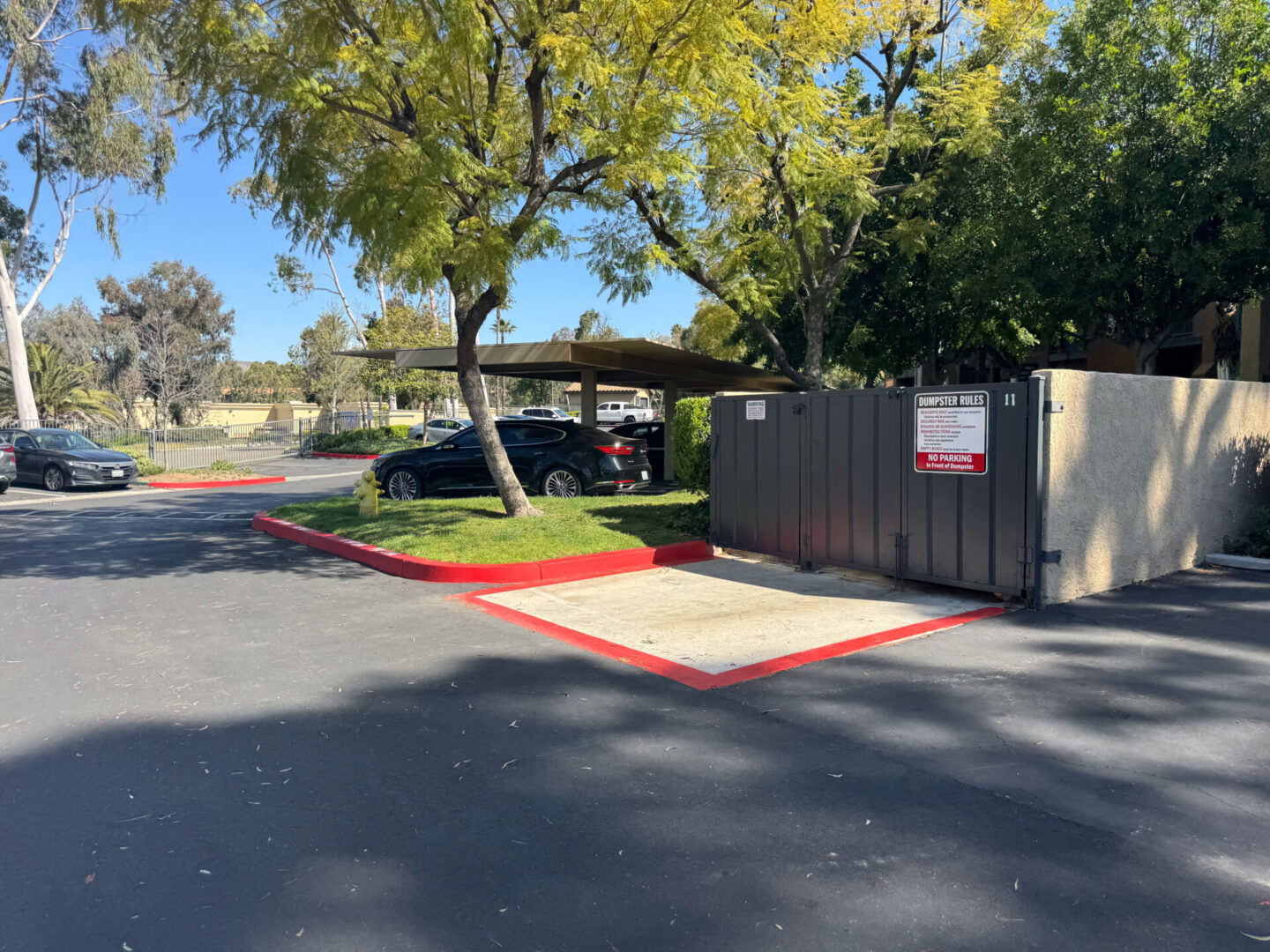 An outdoor parking area with a few cars, bordered by trees and shrubs. A sign on a fenced enclosure with waste bins is visible. The ground is paved, with red-painted curb lines.