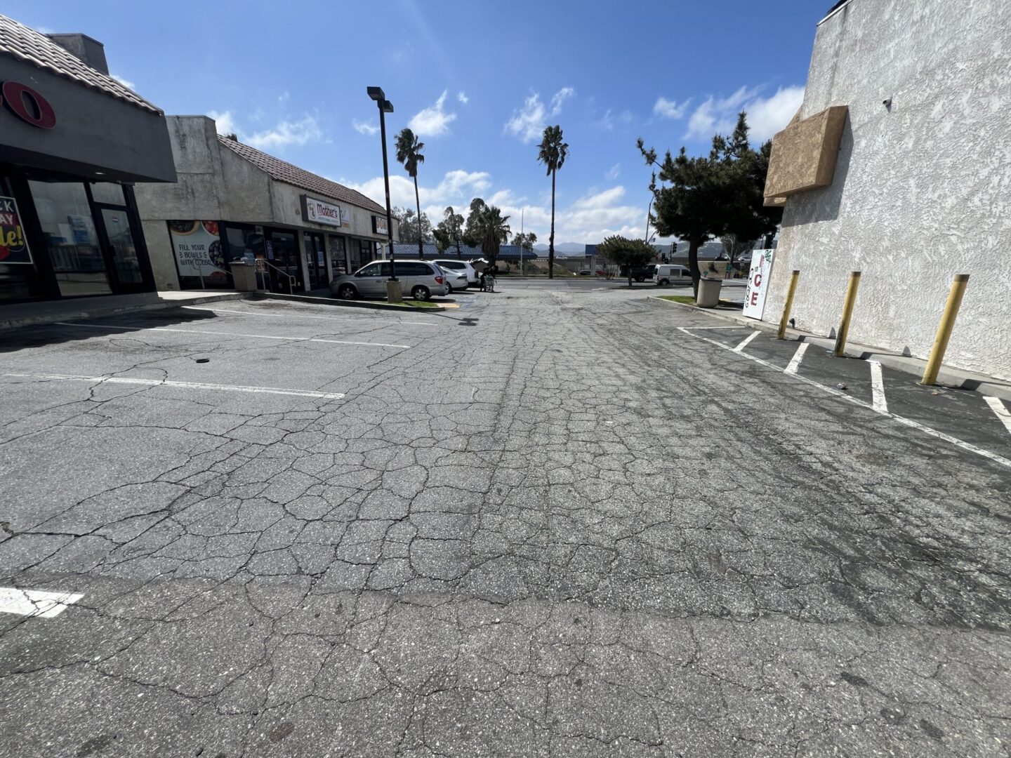 An empty parking lot outside a strip mall with parked cars on the left. The sky is partly cloudy with a few palm trees visible in the background.