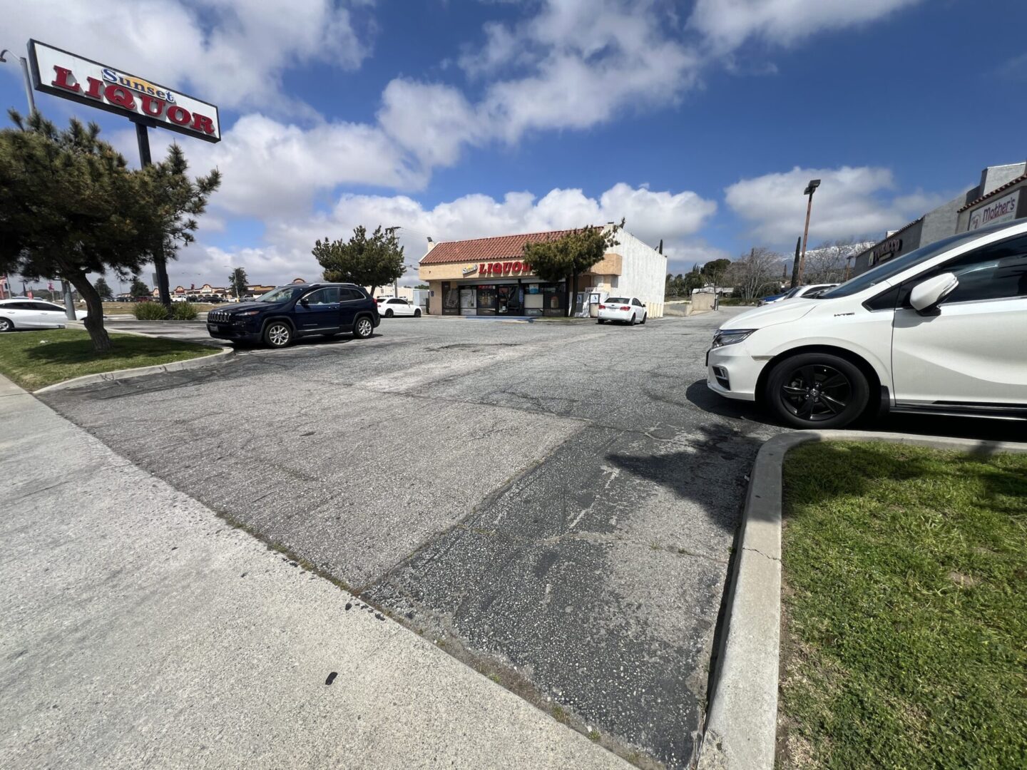 A liquor store with a large sign is located in a small shopping plaza. Several cars are parked in the lot, and the sky is partly cloudy.