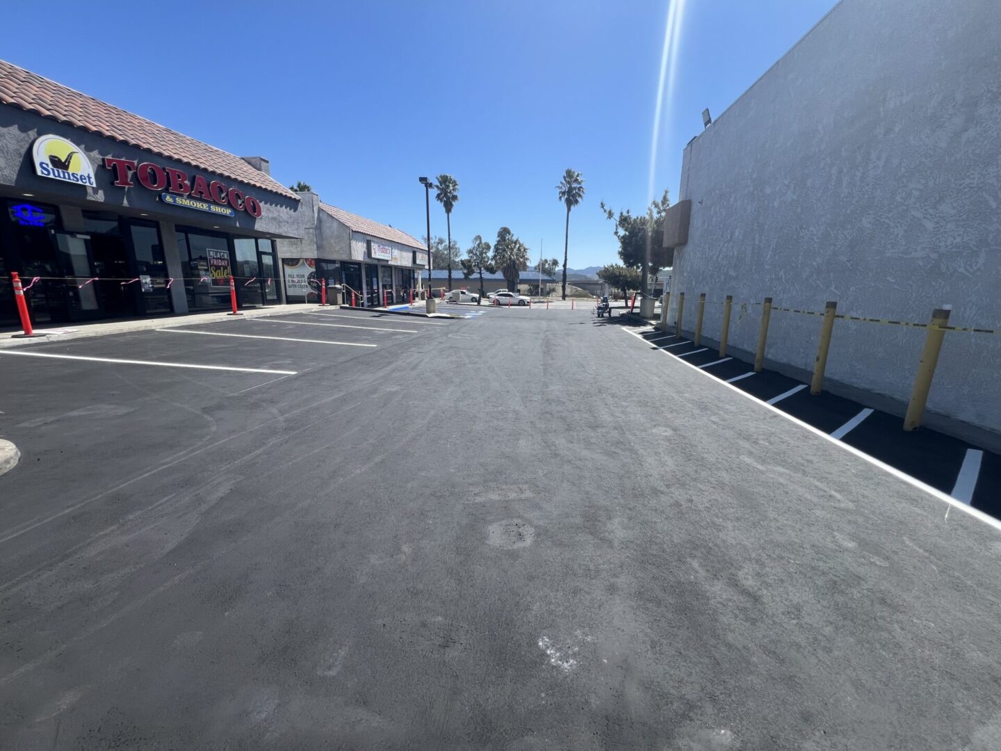 A newly paved parking lot in front of a tobacco shop under a clear blue sky, with empty parking spaces, yellow poles on the right side, and distant palm trees on the horizon.