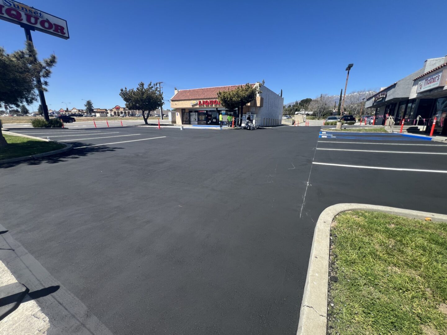 A freshly paved, empty parking lot in front of a liquor store and other small shops on a sunny day.